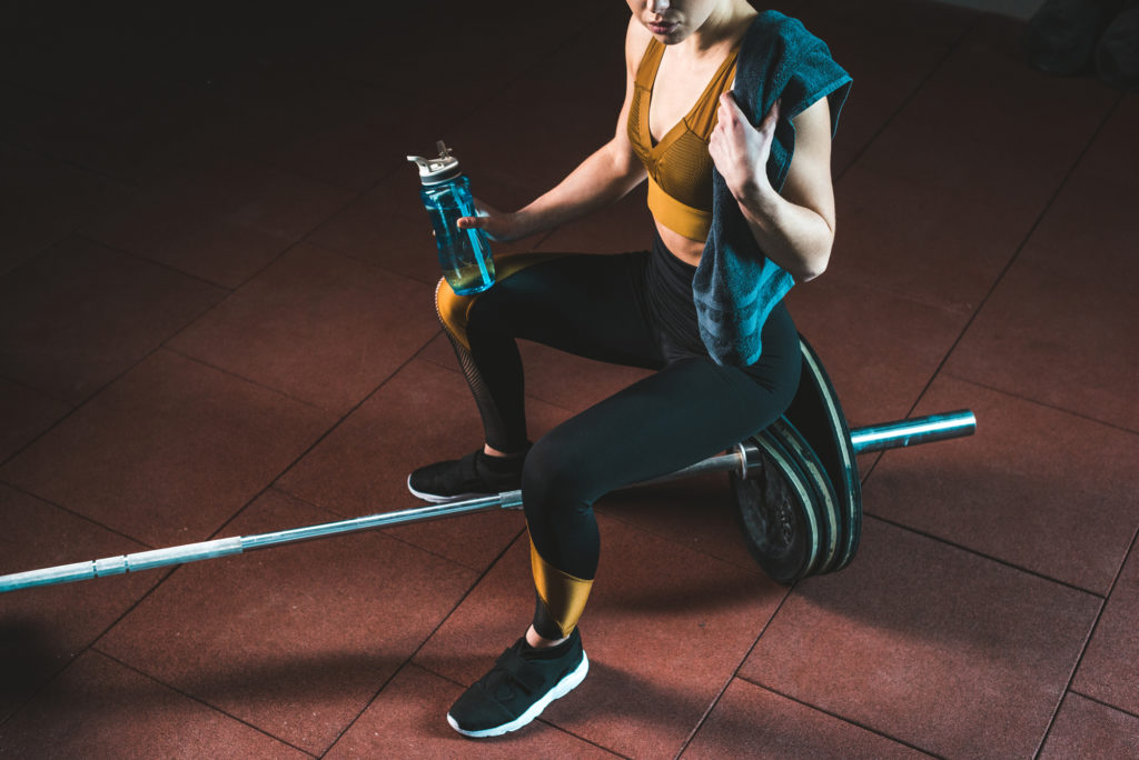 Woman sitting on weights, taking a break from working with a Body building trainer Austin, TX while holding a towel and water bottle